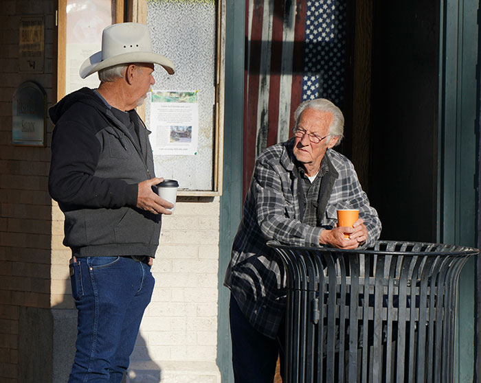 Two older men having a casual conversation outside, one holding a cup, both involved in a relaxed interaction.