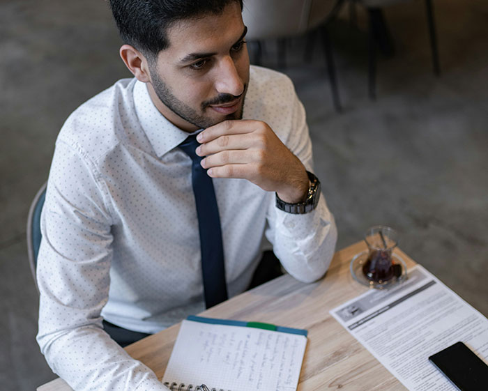 A man in a shirt and tie, holding a pen and sitting at a desk with documents, casually reflecting.