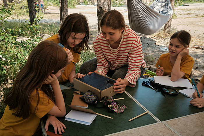 Children outdoors, focused on crafting at a table, surrounded by trees, illustrating an interesting coincidence happening.