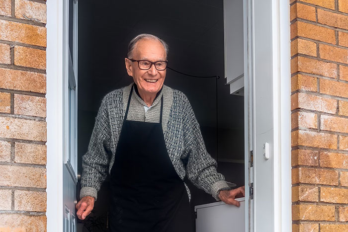 Elderly man with glasses smiles warmly, standing in a doorway.