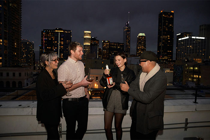 Group of four friends on a rooftop at night, raising glasses, with a city skyline background.