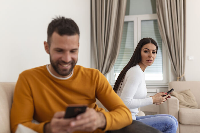 A couple sitting apart; woman looks concerned while man smiles at his phone, highlighting relationship challenges.