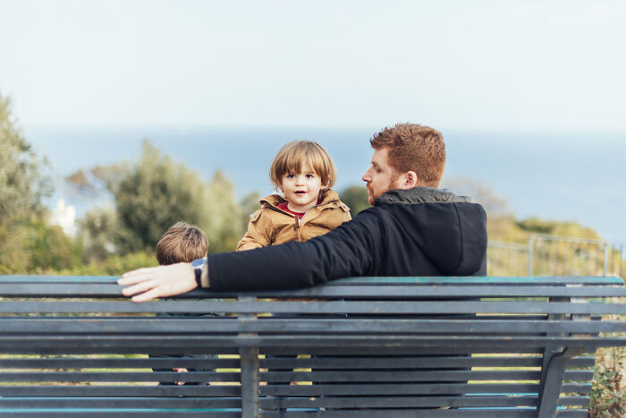 A man and two children sitting on a park bench with a scenic ocean view, highlighting family togetherness in hard times.