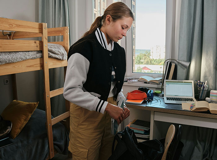 Student organizing items in a college dorm room with a loft bed and study desk visible.