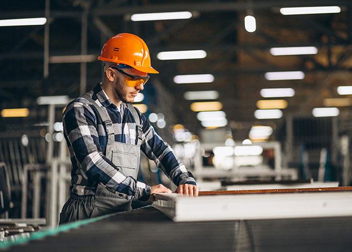 A worker in a plaid shirt and orange helmet operates machinery indoors, emphasizing safety precautions.