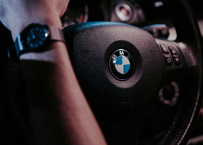 Hand on BMW steering wheel, symbolizing ignoring advice despite warnings; focus on branding and risky decisions.