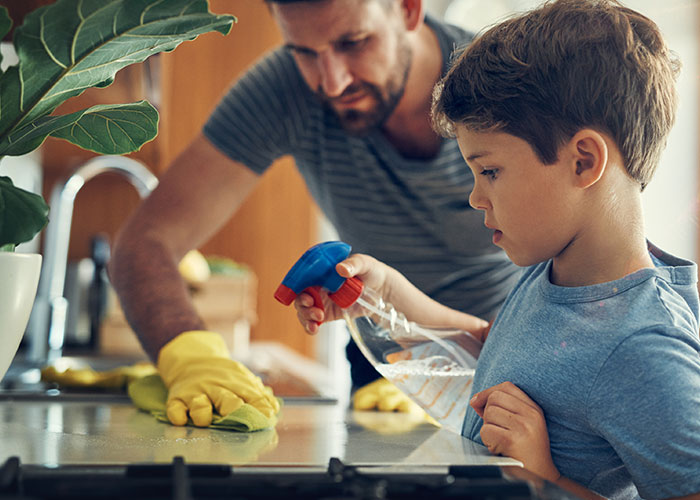 Father and son cleaning a kitchen counter together, using spray bottles and gloves.