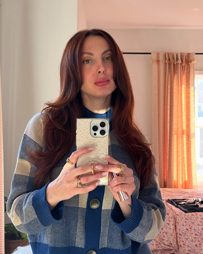 A woman with red hair taking a mirror selfie, wearing a blue and beige striped cardigan.