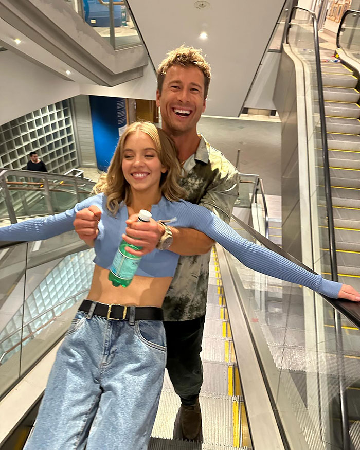 Sydney Sweeney smiling playfully on an escalator, wearing casual attire, causing fan debates about her dress choice.
