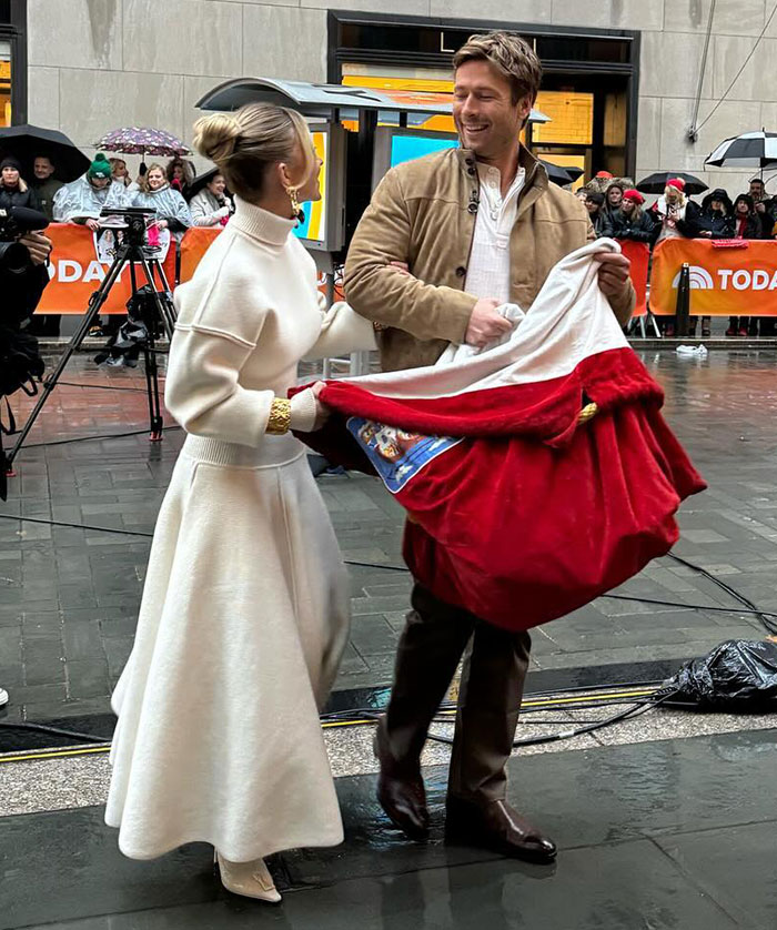 Sydney Sweeney in white dress discusses with man in beige jacket, surrounded by onlookers and camera crew.