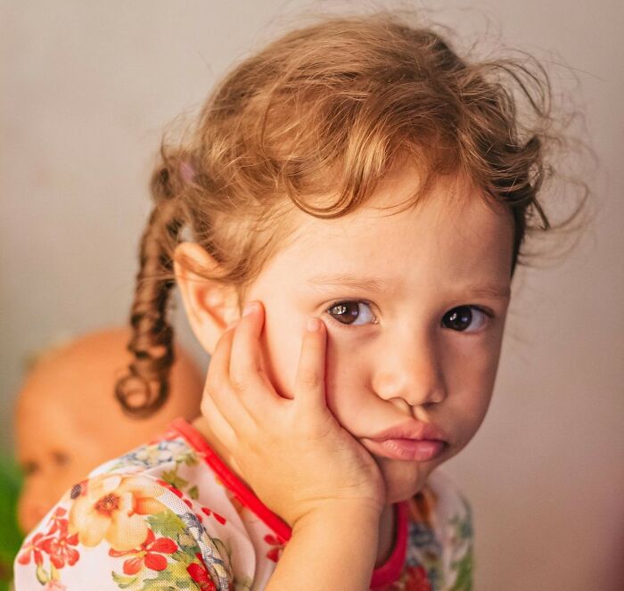 Young child with curly hair and floral shirt, resting face on hand, looking pensive in soft lighting.