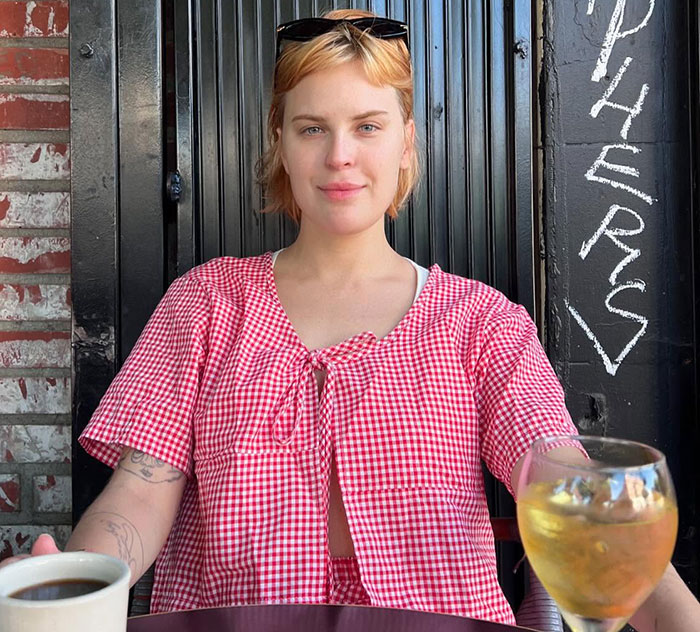 Young woman in red checkered shirt sitting outdoors with coffee and wine, suggesting a comment about Ashton Kutcher.