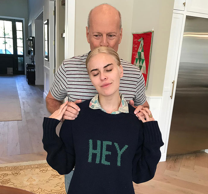 Demi Moore's daughter in a navy sweater with "HEY," standing with eyes closed, someone behind her indoors.