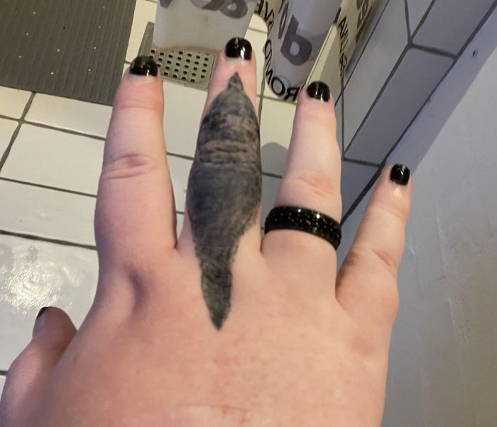 Black ink tattoo fail on finger resembling a distorted shape with black-painted nails and a black ring.