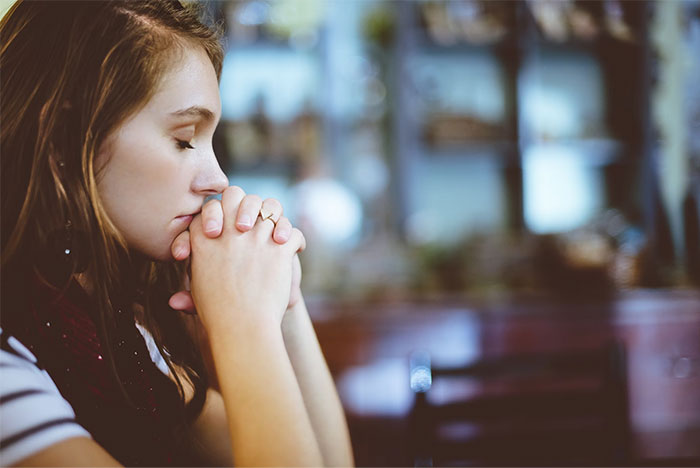 A thoughtful woman with closed eyes, hands clasped, reflecting silently in a dimly lit room.