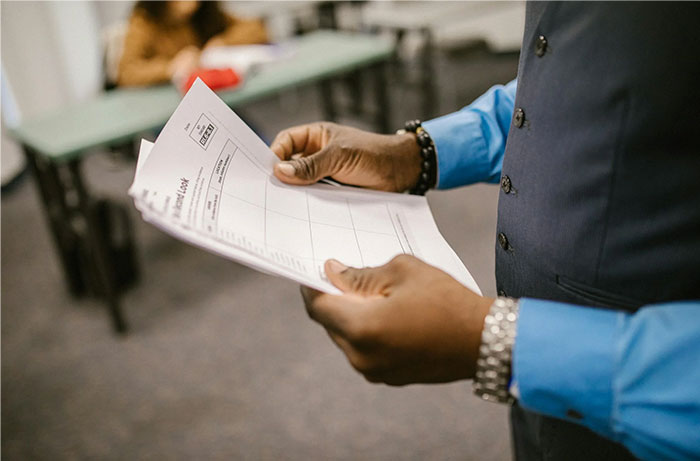 Teacher holding papers in a classroom, discussing authority and student perceptions.