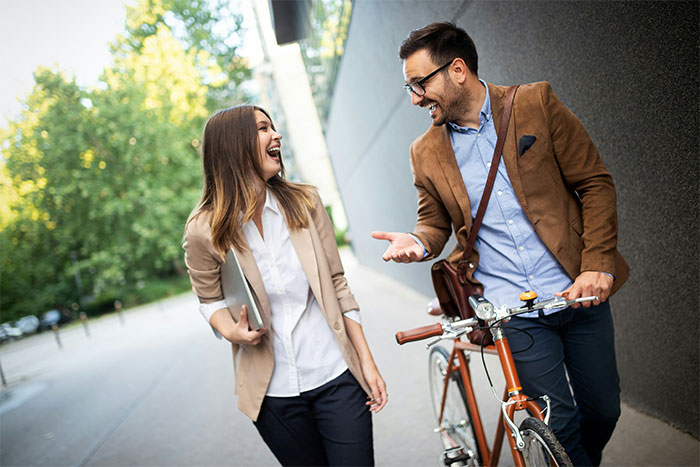 Two teachers in casual conversation, carrying a laptop and bicycle, laughing while walking outdoors.