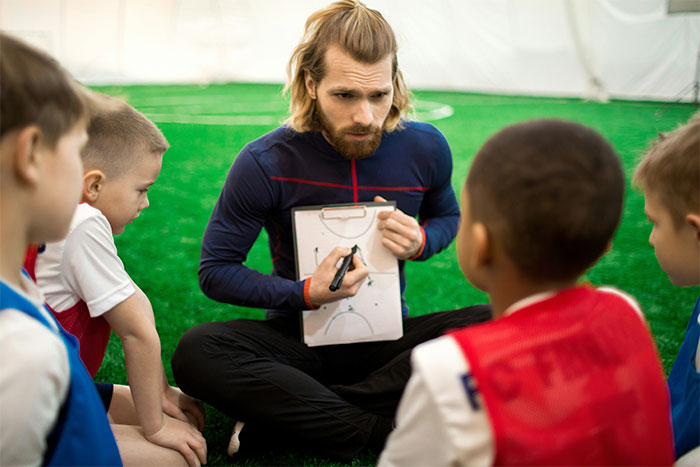 Teacher coaching young students on a field, diagramming strategies, maintaining authority and focus indoors.