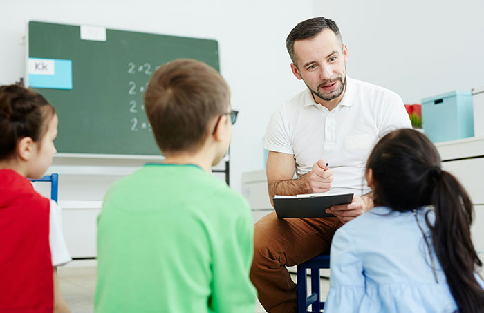 Teacher with clipboard talking to students in a classroom, green chalkboard in background, symbolizing authority dynamics.