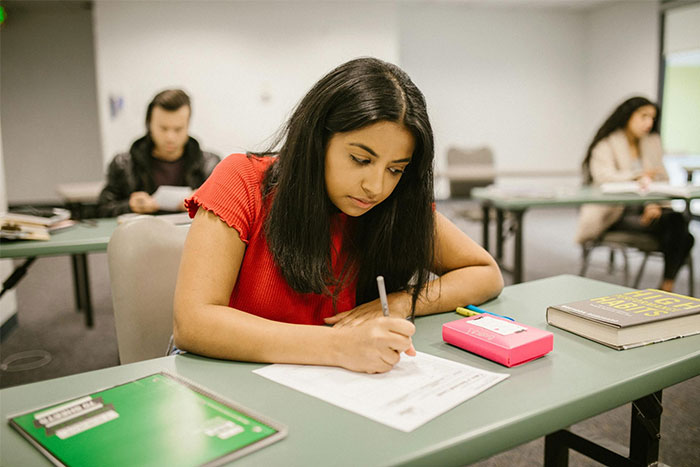 Student in a red shirt focused on writing an exam in a classroom setting, with school supplies on the desk.