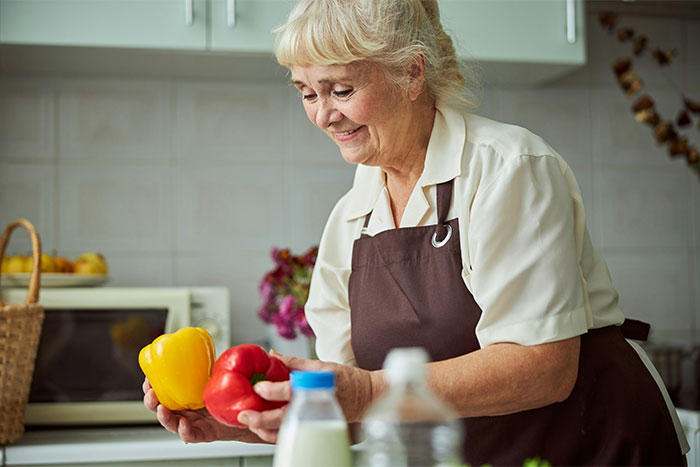 Elderly woman in a kitchen holding peppers, wearing a brown apron and smiling.