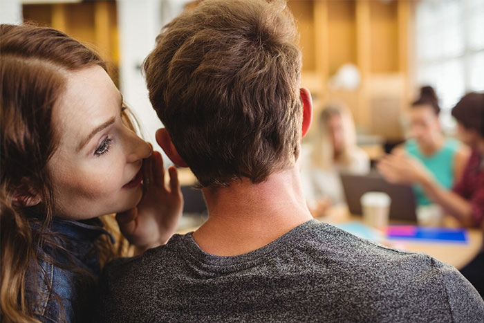 Teacher whispering a secret to a student in a classroom setting, with students in the background.