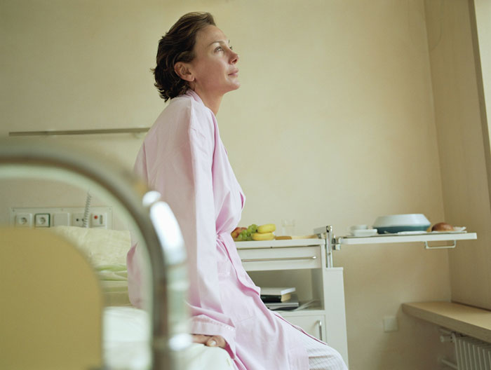 Woman in a pink robe sitting on a hospital bed, gazing thoughtfully, reflecting on an engagement dinner.