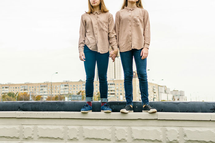 Two people in matching outfits holding hands, standing on a ledge, with buildings in the background, evoking an uncomfortable mood.