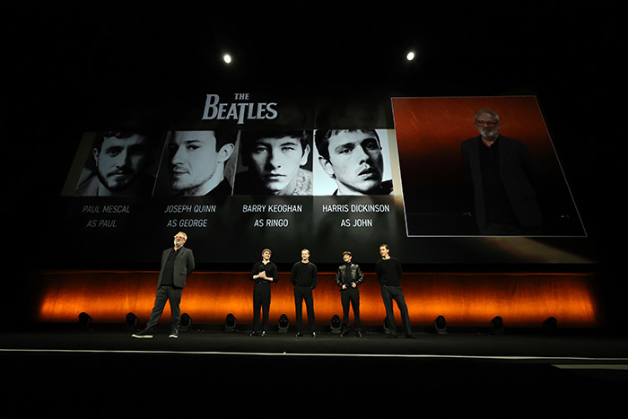 Casting announcement for The Beatles biopic, featuring actors on stage with projected images of their roles.