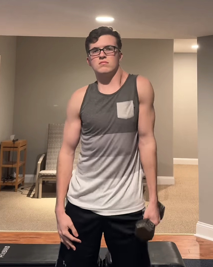 Young man in a gym wearing glasses and a tank top, holding a dumbbell in one hand, highlighting uneven workout results.
