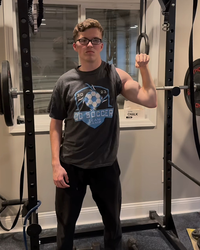 Young man in glasses showcasing his right arm's muscle development, standing near workout equipment, "The Crooked Man.