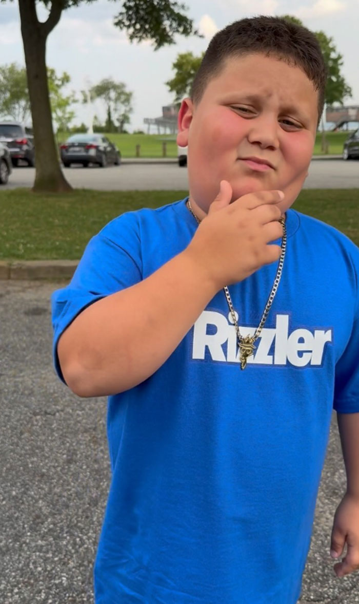 The Rizzler wearing a blue Rizzler shirt and necklace, posing with hand on chin in outdoor parking lot setting.