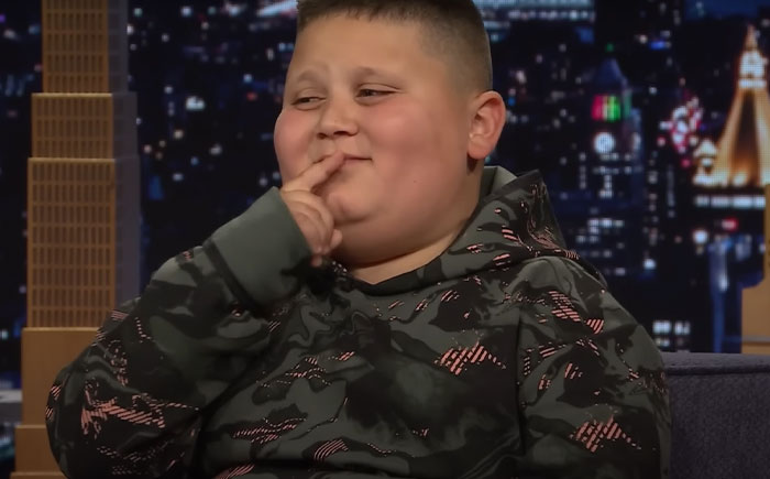 Young boy known as the Rizzler smiling on a late-night talk show set with cityscape backdrop behind him