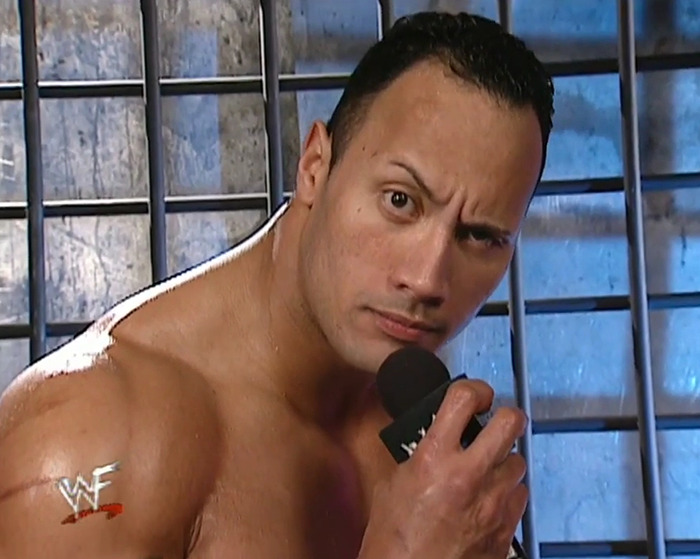 Dwayne Johnson in classic wrestling setting holding a microphone, sparking new movie race confusion.