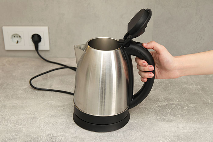 Hand holding a European electric kettle with an open lid on a kitchen counter.