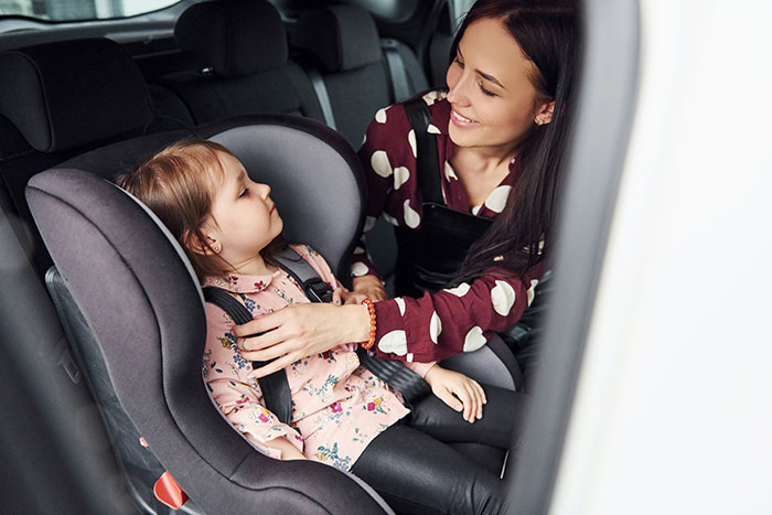 A woman secures a child in a car seat, smiling inside a vehicle.