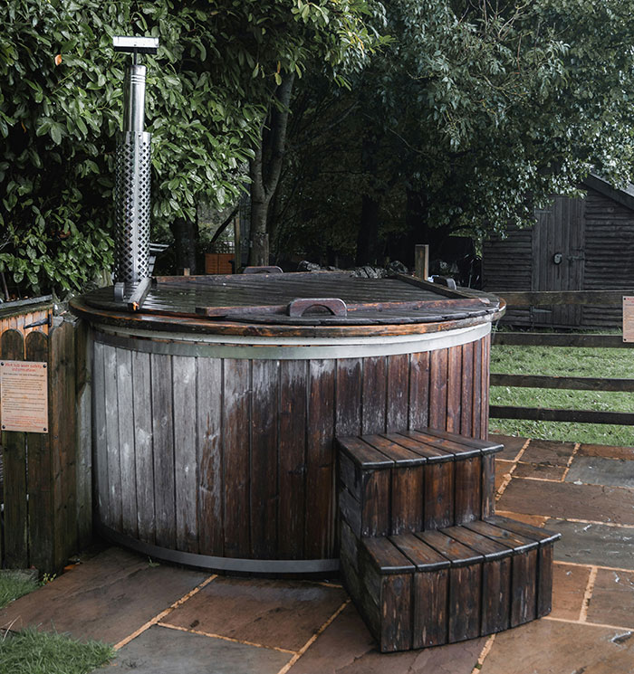 Wooden outdoor hot tub with steps in a garden setting, surrounded by greenery.