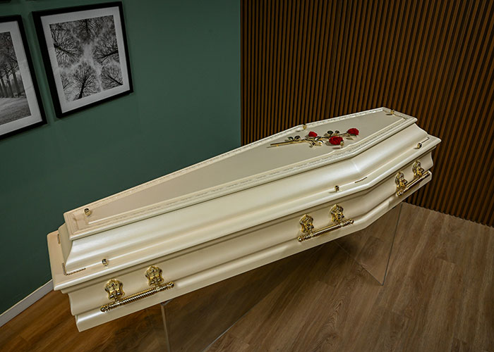 Elegant white coffin in a room with wooden walls, showing red roses on top and framed photographs in the background.