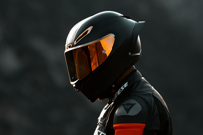 A motorcyclist wearing a sleek black motorcycle helmet with an orange visor against a blurred background.
