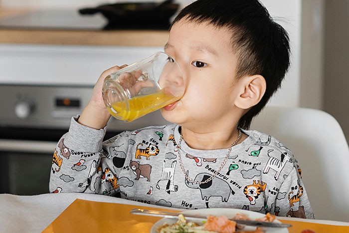 Child drinking juice at home for safety, wearing a printed sweatshirt, sitting at a dining table with food.