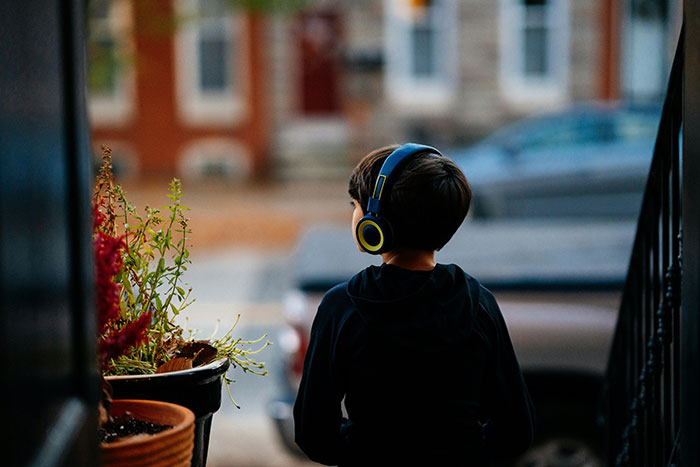 Child wearing headphones, looking out a doorway onto a street, symbolizing life-changing precautions.
