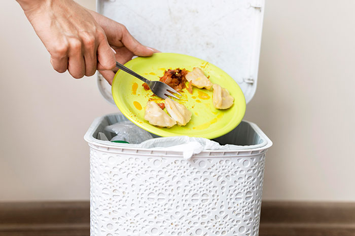 Hand discarding food from a yellow plate into a white trash can. Don't forget to clean before hosting guests.