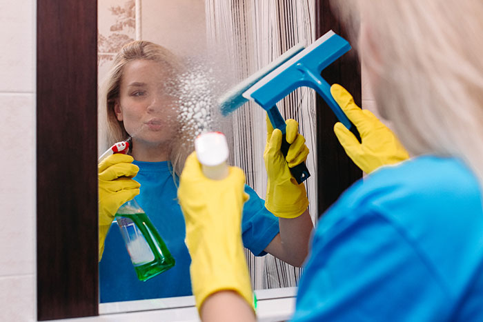 Person cleaning mirror with spray and squeegee, wearing gloves, highlighting cleaning tips before hosting guests.