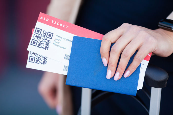 Hand holding an affordable flights ticket and blue passport, resting on a suitcase handle.