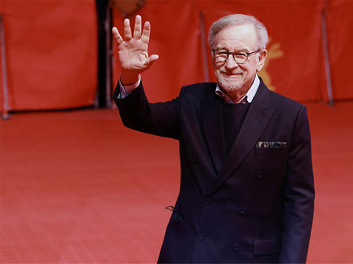 A person in a black suit waving against a red background at a public event.