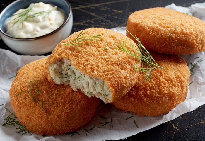 Golden breaded fish cakes garnished with fresh dill, served alongside a bowl of creamy tartar sauce on parchment paper.