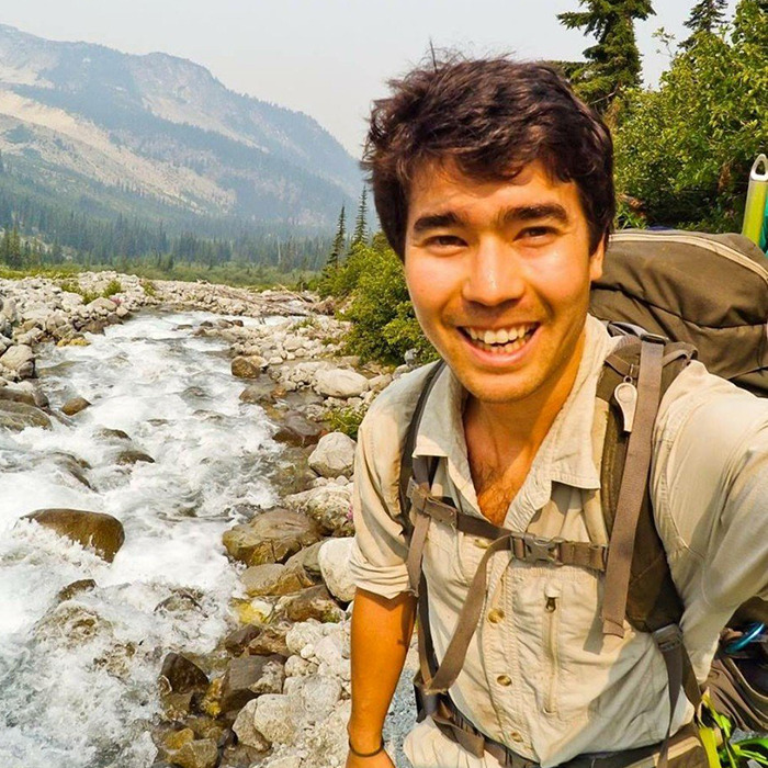 Smiling YouTuber on a mountain trail with river in background, carrying a backpack.