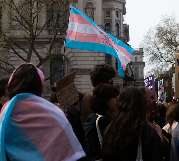 Protest with trans pride flag, people holding signs, highlighting school suspension transphobia issue.