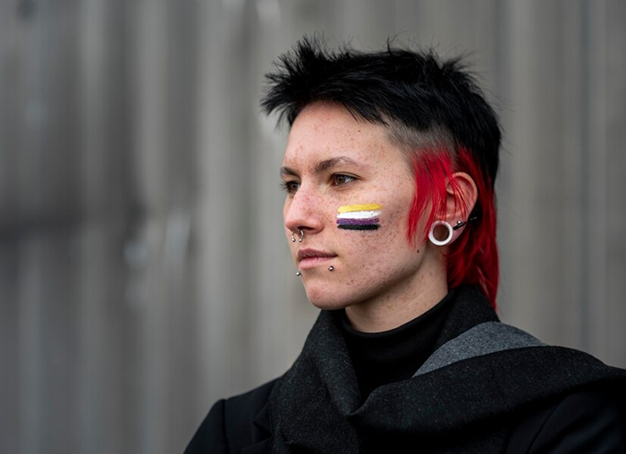 Person with short black and red hair and nonbinary pride flag face paint, representing themes of trans identity and self-discovery. Person with short black and red hair and nonbinary pride flag face paint, representing themes of trans identity and self-discovery.