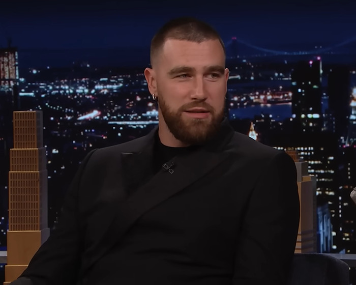 Travis Kelce in a black suit on a talk show with a cityscape background. Travis Kelce in a black suit on a talk show with a cityscape background.
